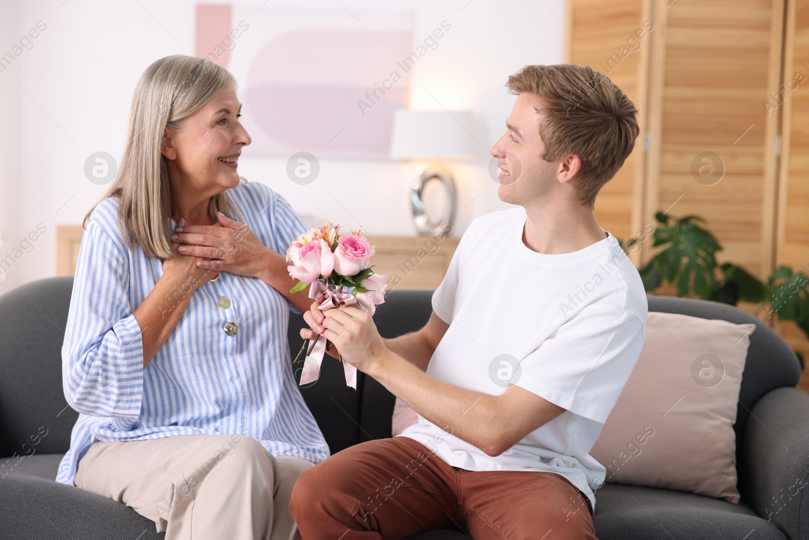 Photo of Son giving his happy mother bouquet of flowers on sofa at home