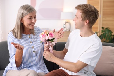 Photo of Son giving his happy mother bouquet of flowers on sofa at home