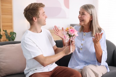 Photo of Son giving his happy mother bouquet of flowers on sofa at home