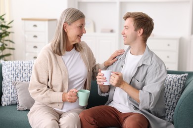 Photo of Happy son and his mother with cups spending time together on sofa at home