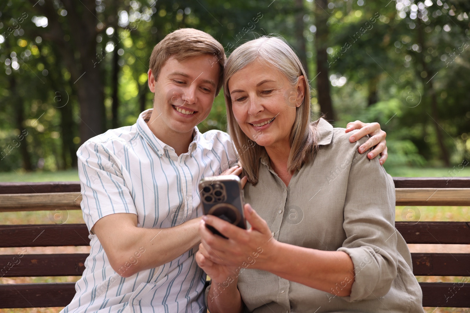 Photo of Smiling mother and her son using smartphone on bench in park