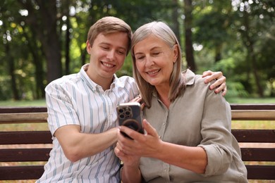 Photo of Smiling mother and her son using smartphone on bench in park