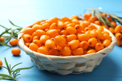 Photo of Fresh sea buckthorn berries in wicker bowl and leaves on light blue wooden table, closeup