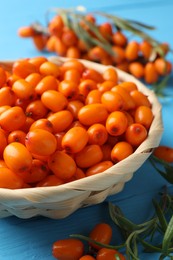 Photo of Fresh sea buckthorn berries in wicker bowl and leaves on light blue wooden table, closeup