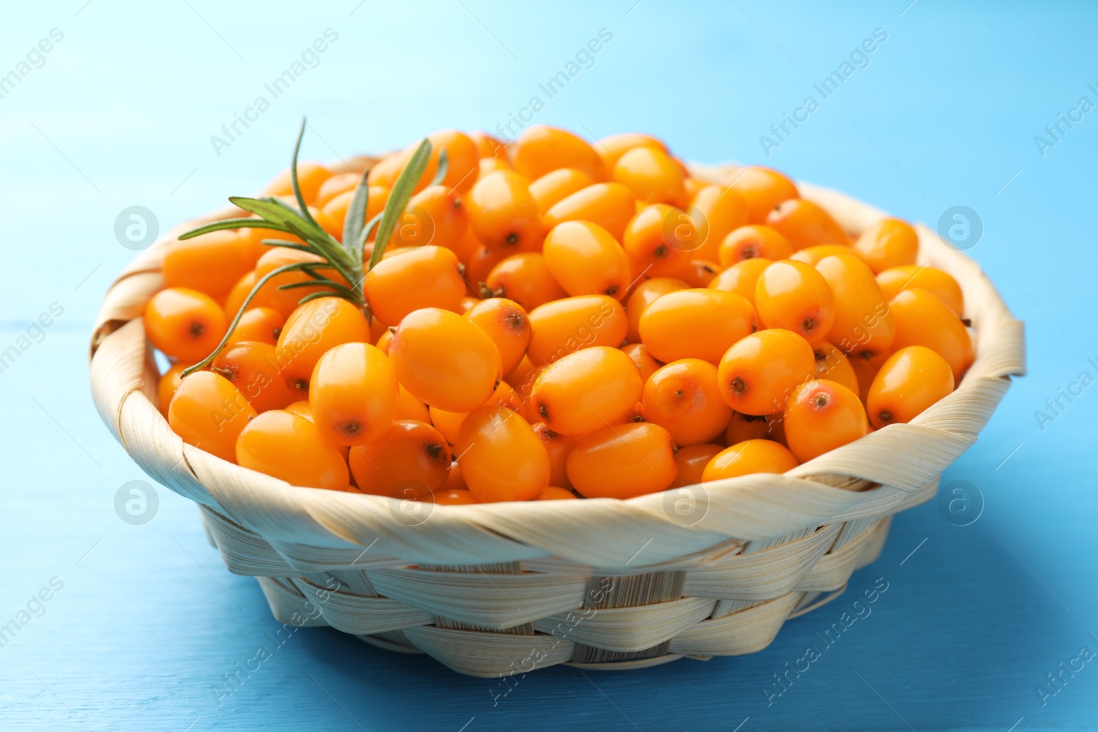 Photo of Fresh sea buckthorn berries in wicker bowl and leaves on light blue wooden table, closeup