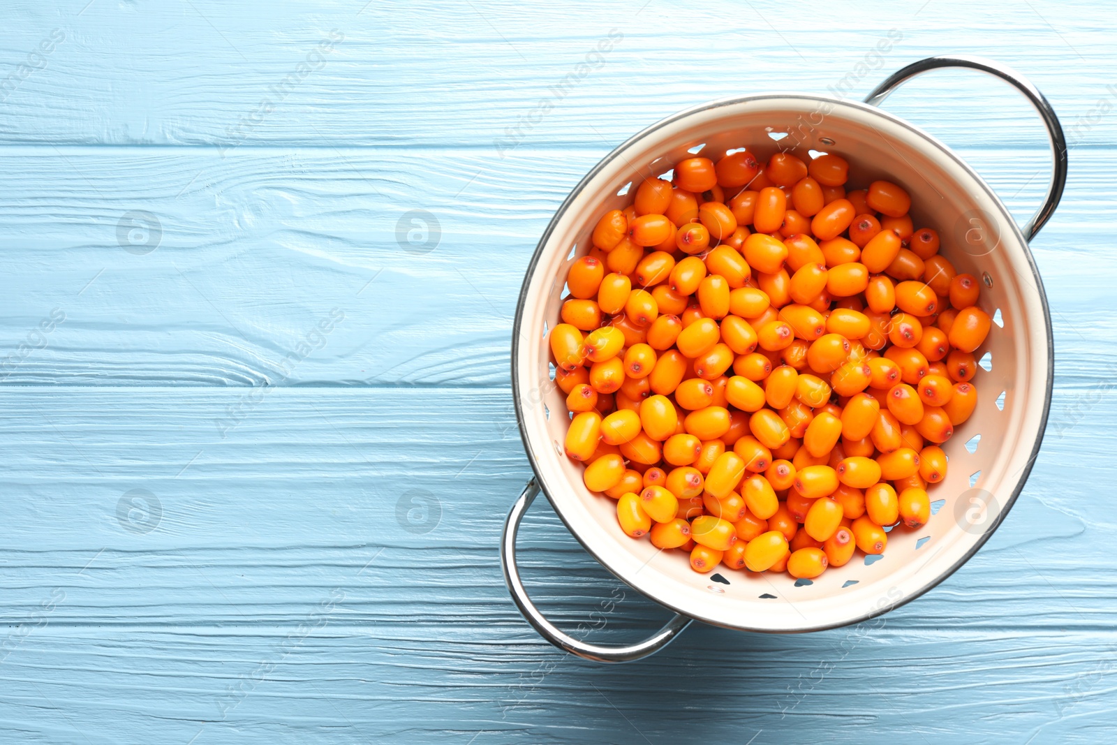 Photo of Fresh sea buckthorn berries in metal colander on light blue wooden table, top view. Space for text