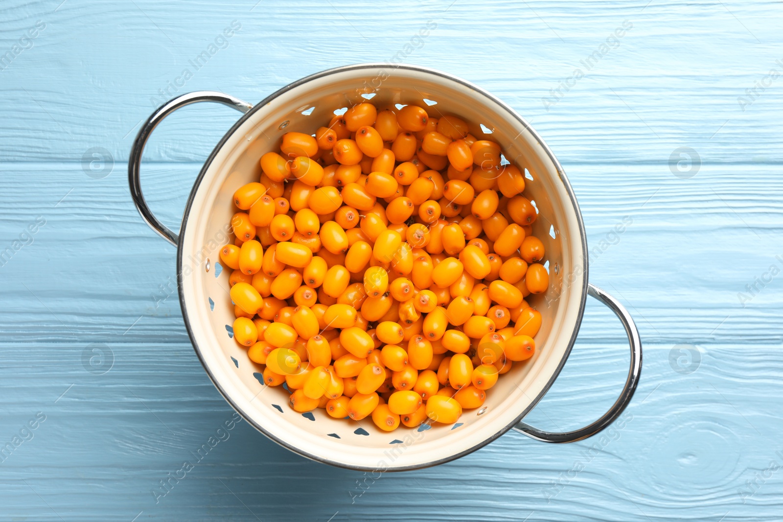 Photo of Fresh sea buckthorn berries in metal colander on light blue wooden table, top view