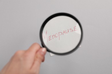 Photo of Woman holding magnifying glass over paper note with word Menopause on light grey background, closeup
