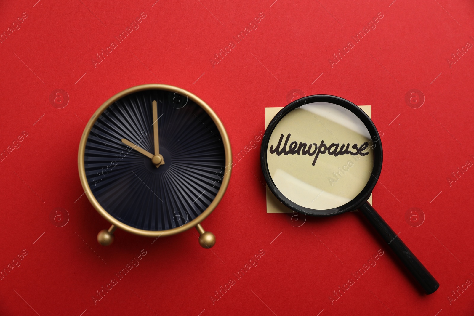 Photo of Magnifying glass over paper note with word Menopause and alarm clock on red background, top view