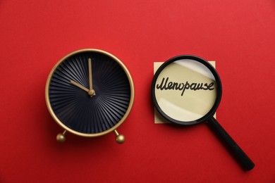 Photo of Magnifying glass over paper note with word Menopause and alarm clock on red background, top view