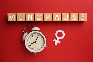 Photo of Wooden cubes with word Menopause, female gender symbol and alarm clock on red background, flat lay
