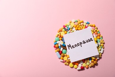 Photo of Paper note with word Menopause and pills on pink background, top view. Space for text