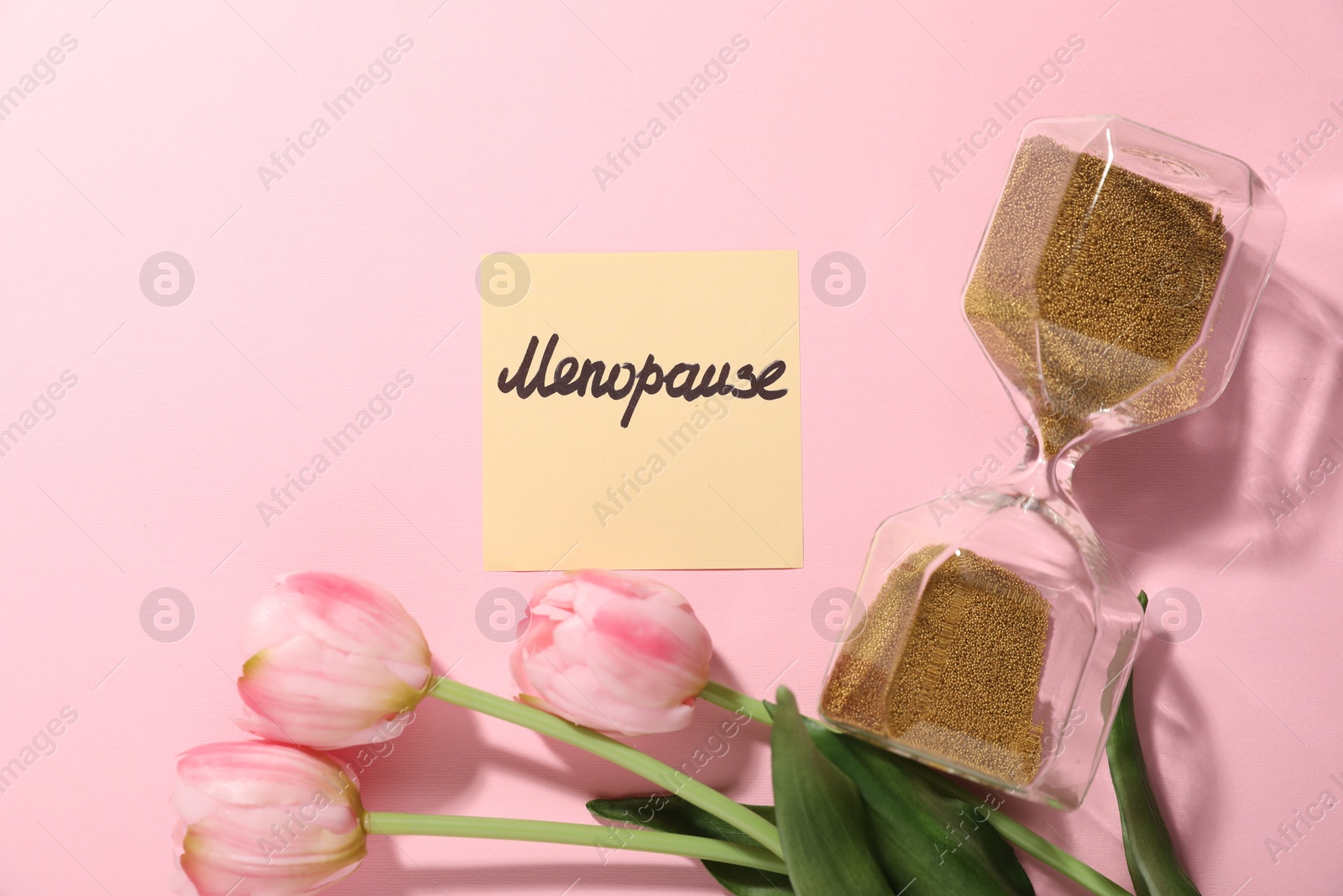 Photo of Paper note with word Menopause, tulips and hourglass on pink background, flat lay