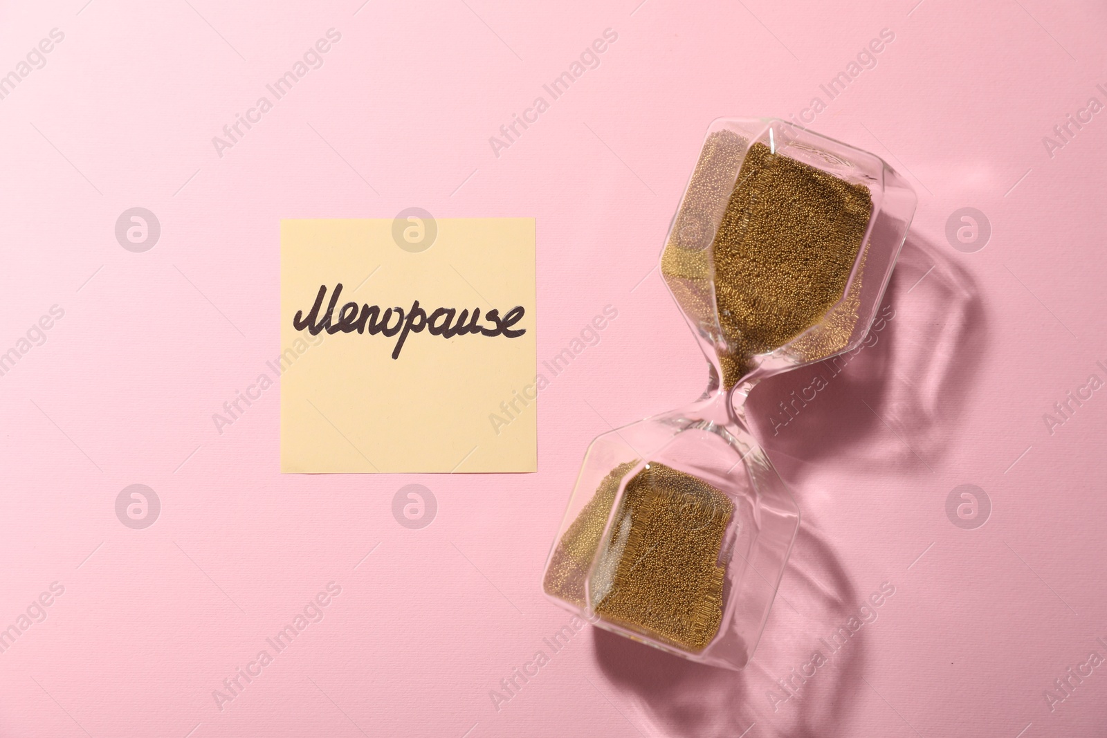Photo of Paper note with word Menopause and hourglass on pink background, top view