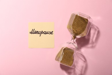 Photo of Paper note with word Menopause and hourglass on pink background, top view