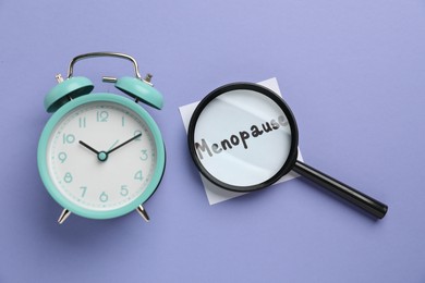 Photo of Magnifying glass over paper note with word Menopause and alarm clock on violet background, top view