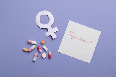 Photo of Paper note with word Menopause, female gender symbol and pills on violet background, flat lay