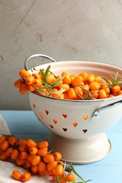 Photo of Fresh sea buckthorn berries in metal colander and leaves on light blue wooden table, closeup
