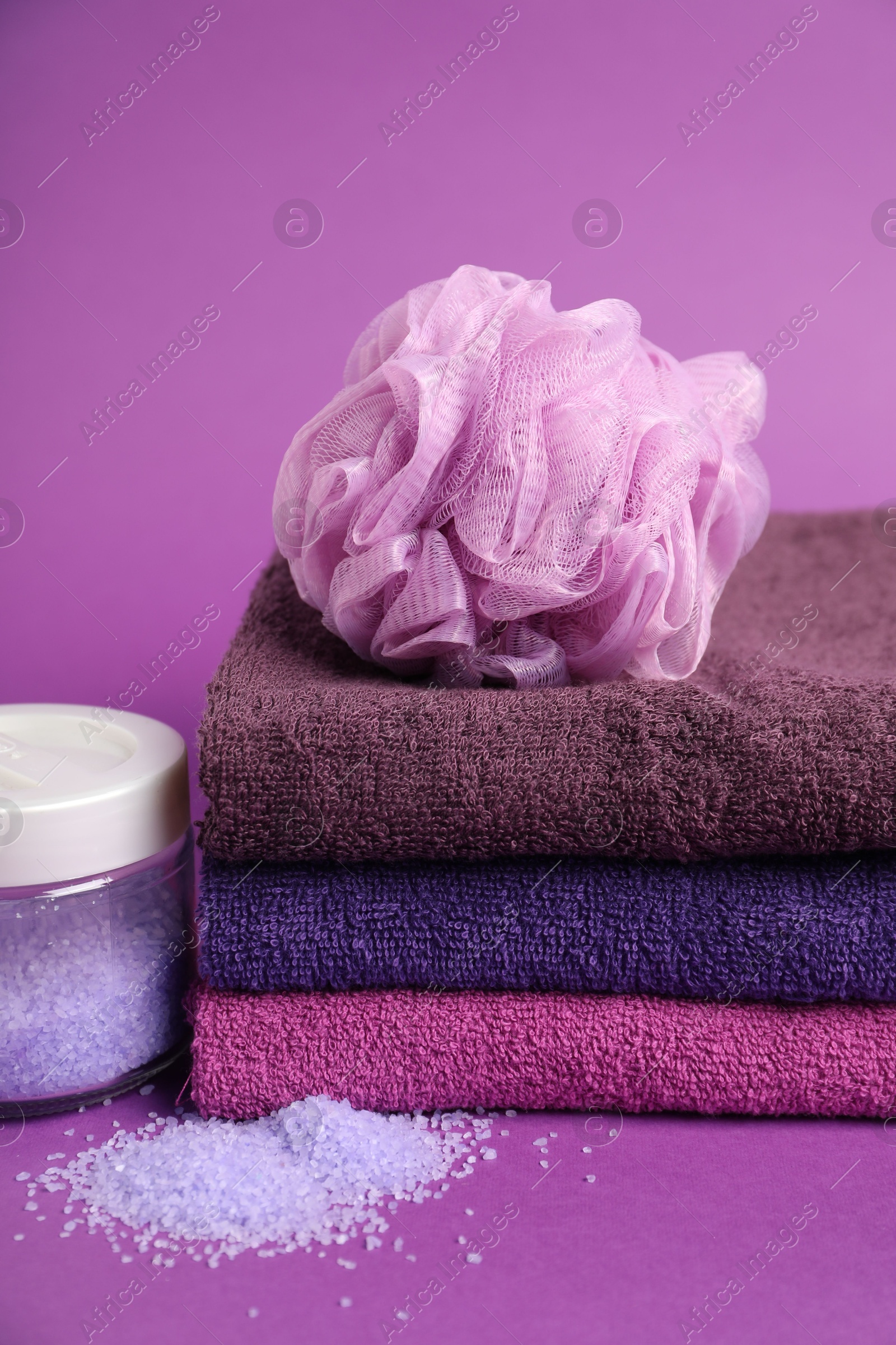 Photo of Stack of fresh terry towels, sponge and sea salt on purple background, closeup