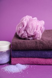 Photo of Stack of fresh terry towels, sponge and sea salt on purple background, closeup