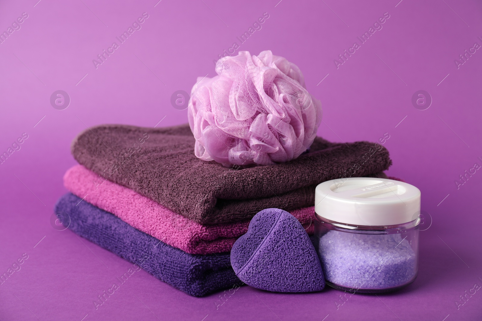 Photo of Stack of fresh terry towels, sponges and sea salt on purple background, closeup
