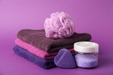 Photo of Stack of fresh terry towels, sponges and sea salt on purple background, closeup