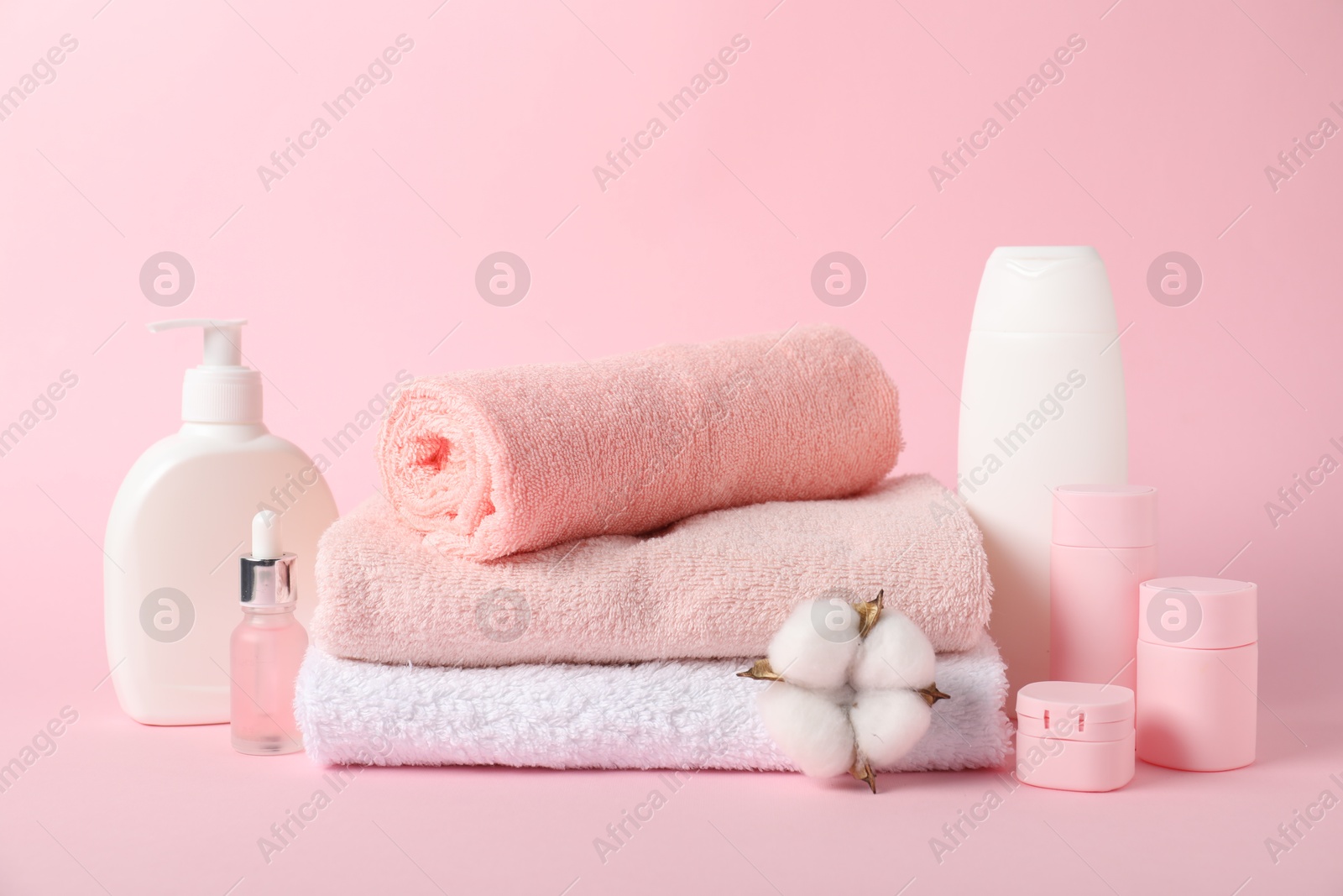 Photo of Stack of fresh terry towels, cosmetic products and cotton flower on pink background