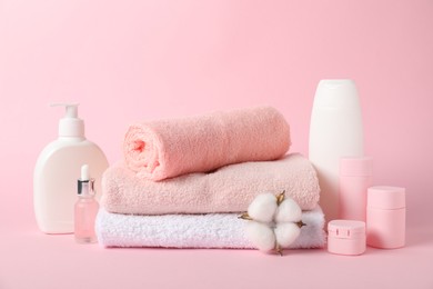 Photo of Stack of fresh terry towels, cosmetic products and cotton flower on pink background