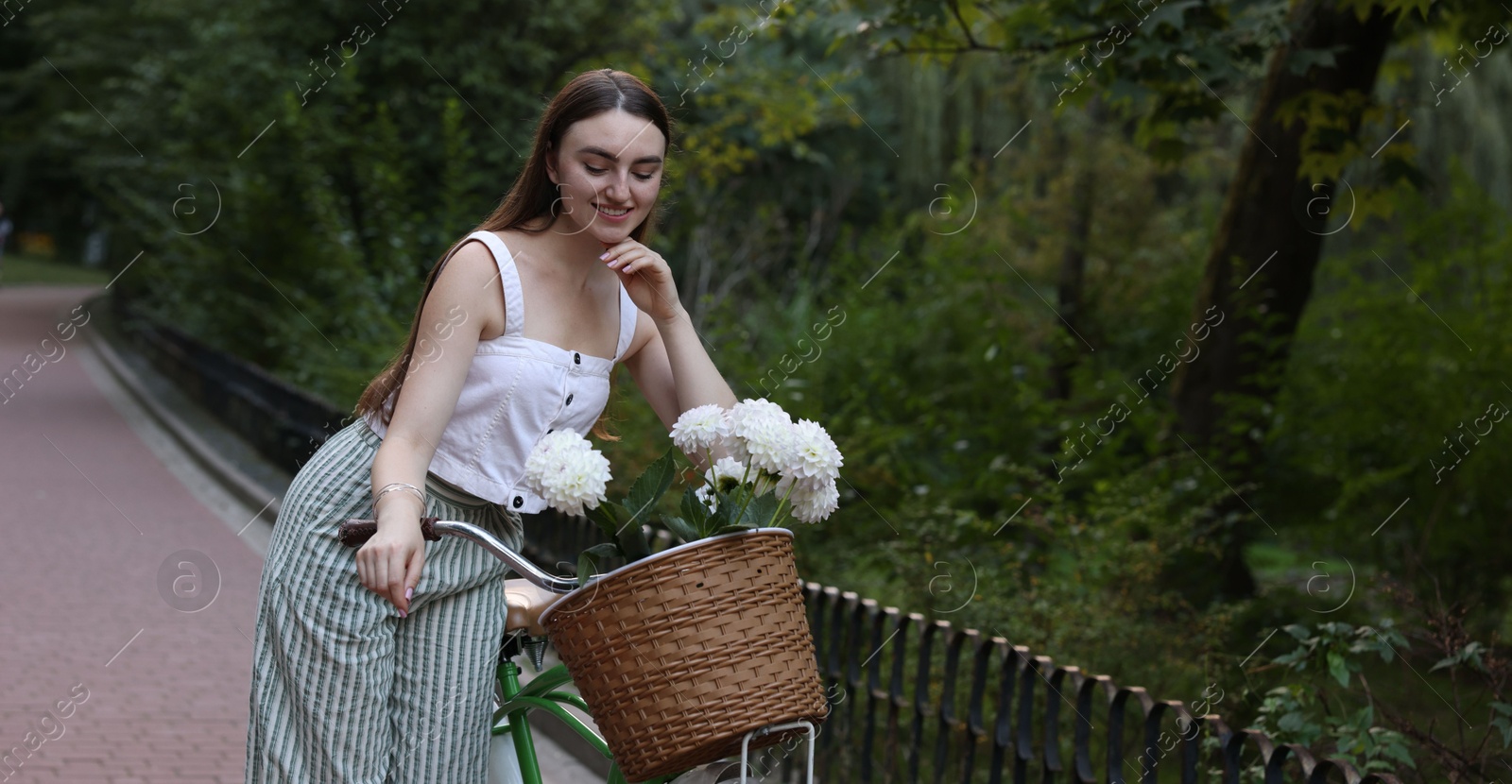 Photo of Woman walking her bicycle with flowers in basket outdoors. Space for text