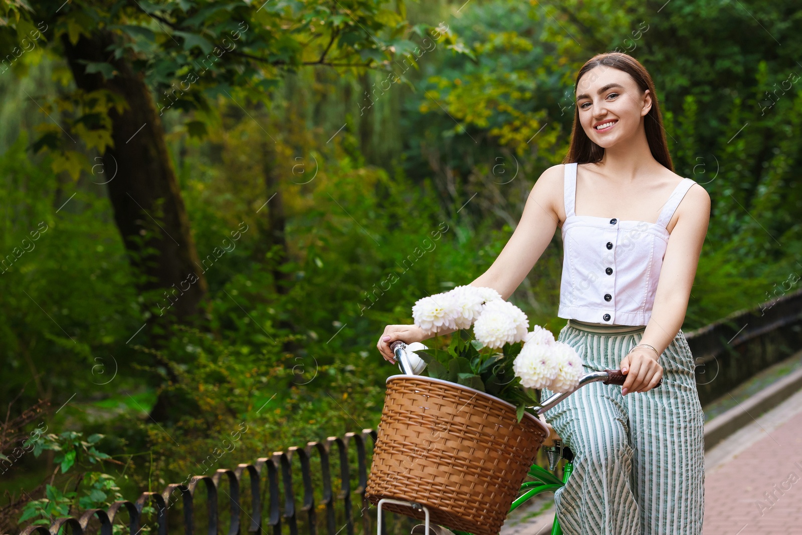 Photo of Woman walking her bicycle with flowers in basket outdoors. Space for text
