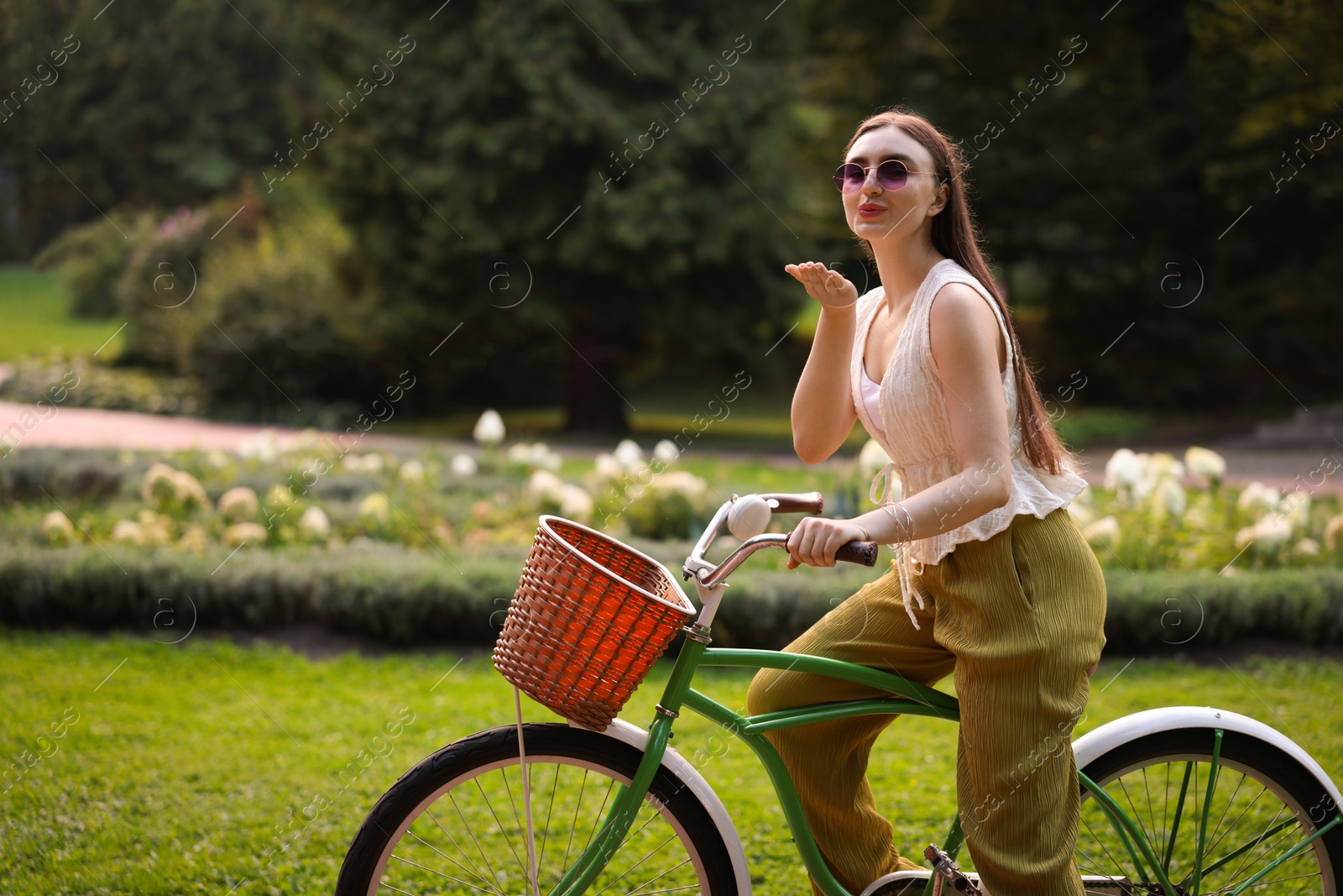 Photo of Happy woman blowing air kiss while riding bicycle in park. Space for text