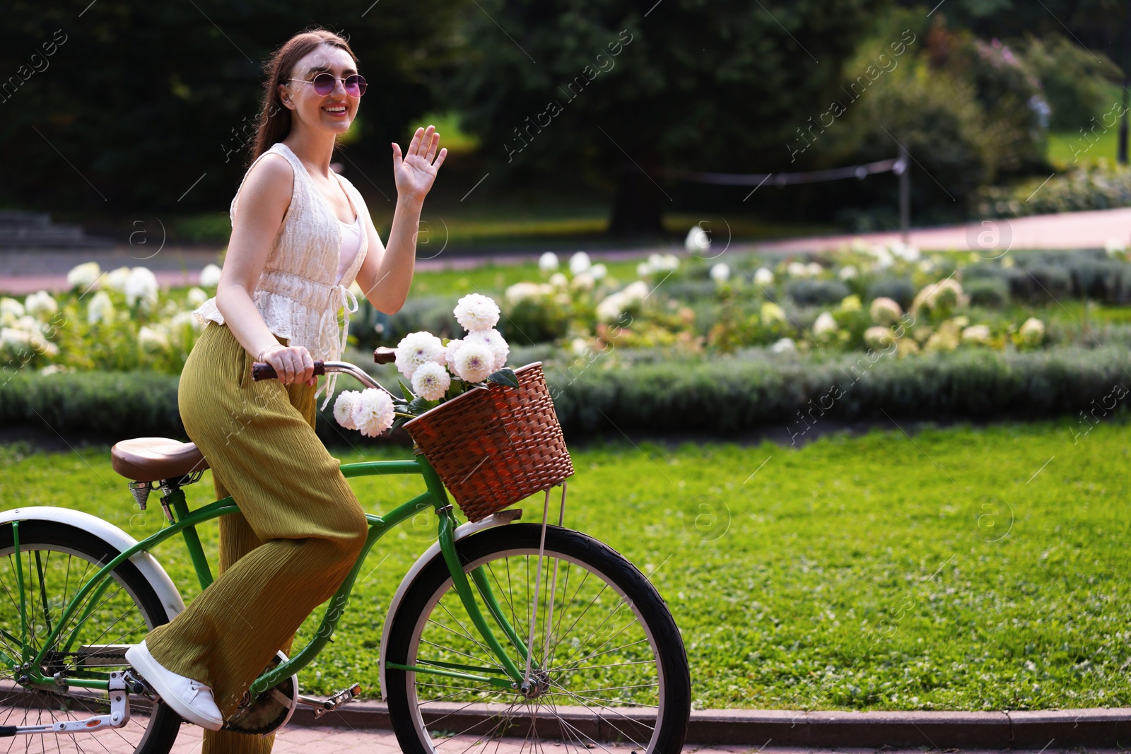 Photo of Woman riding bicycle with flowers in basket and waving outdoors. Space for text