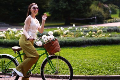 Photo of Woman riding bicycle with flowers in basket and waving outdoors. Space for text