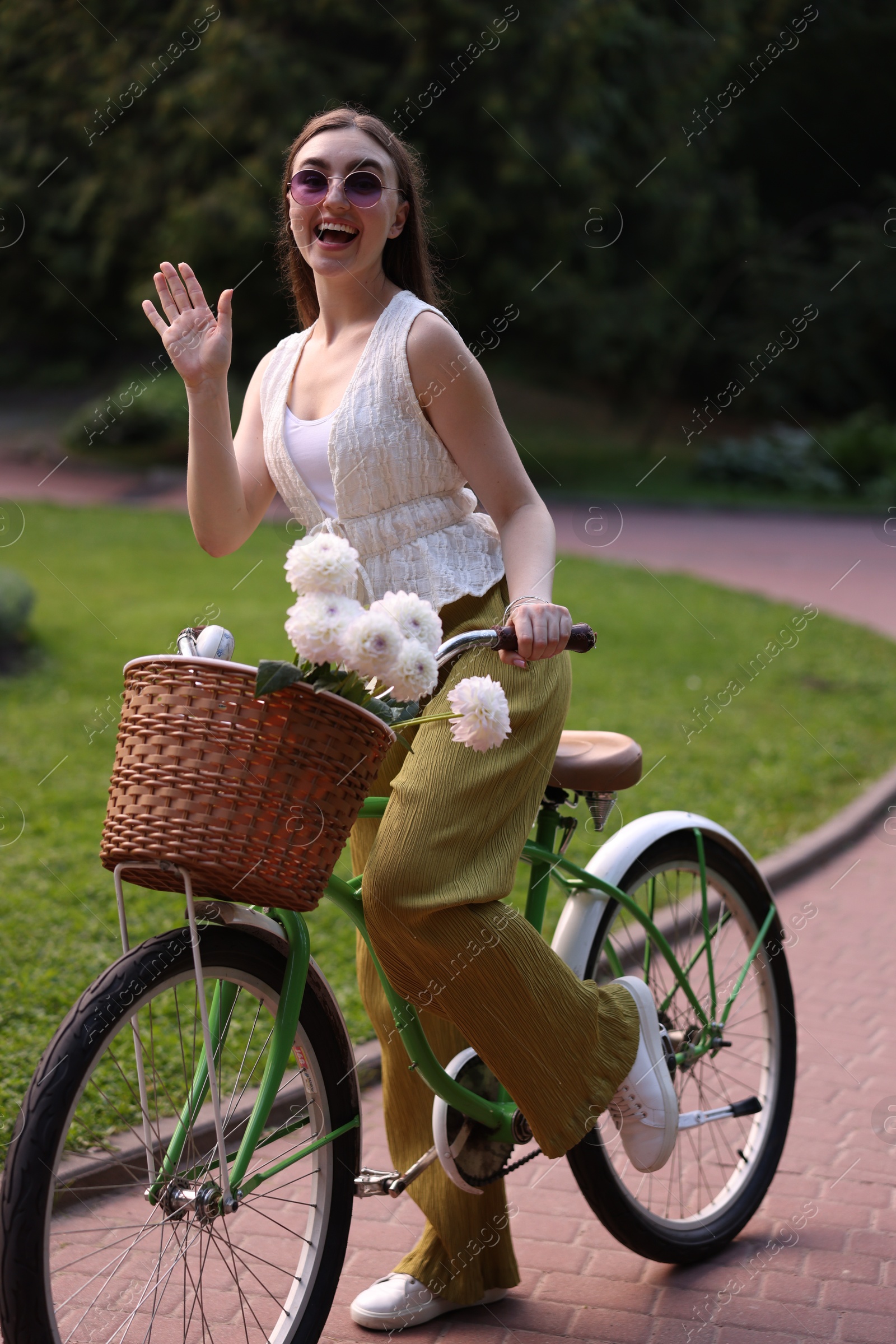 Photo of Woman riding bicycle with flowers in basket and waving outdoors