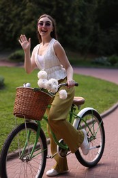 Photo of Woman riding bicycle with flowers in basket and waving outdoors
