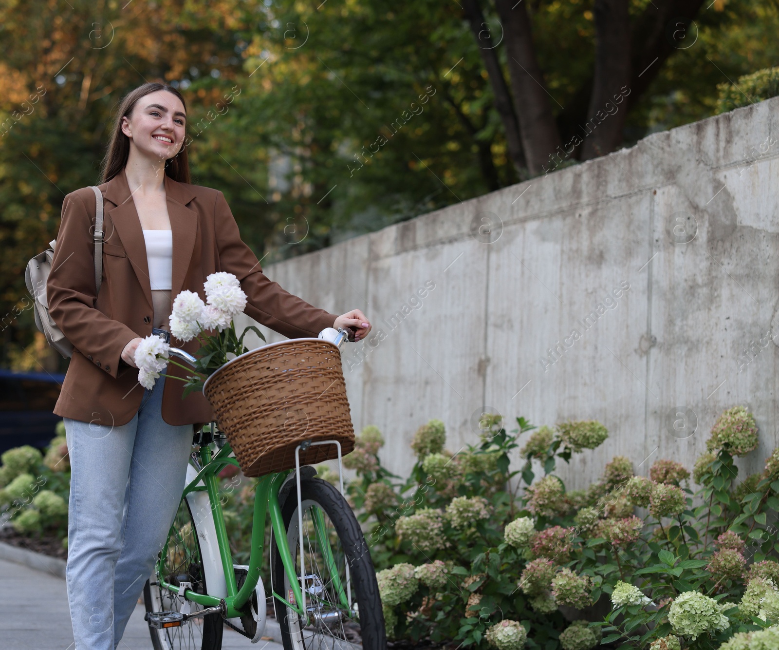 Photo of Woman walking her bicycle with flowers in basket outdoors. Space for text