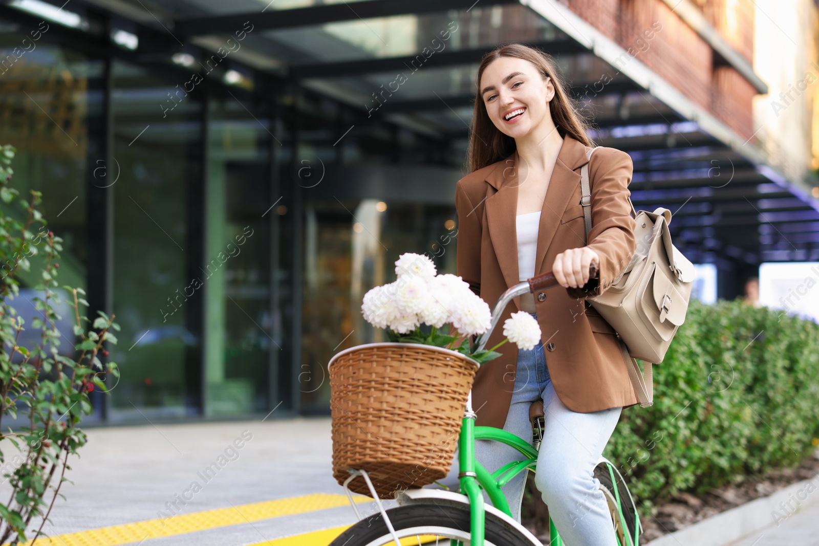 Photo of Woman riding bicycle with flowers in basket on city street. Space for text