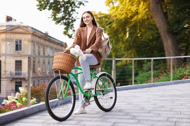 Photo of Woman riding bicycle with flowers in basket on city street. Space for text