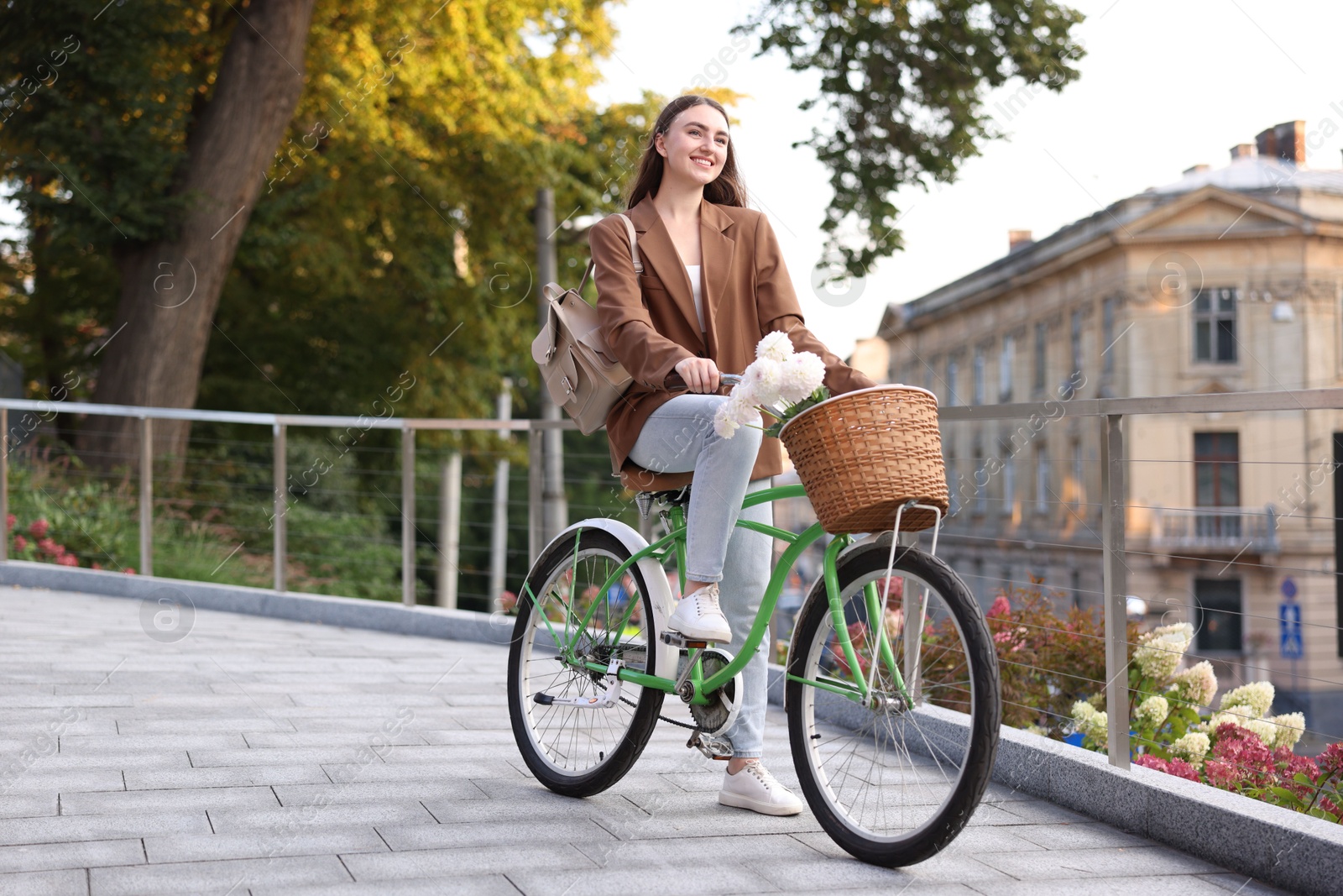 Photo of Woman riding bicycle with flowers in basket on city street. Space for text