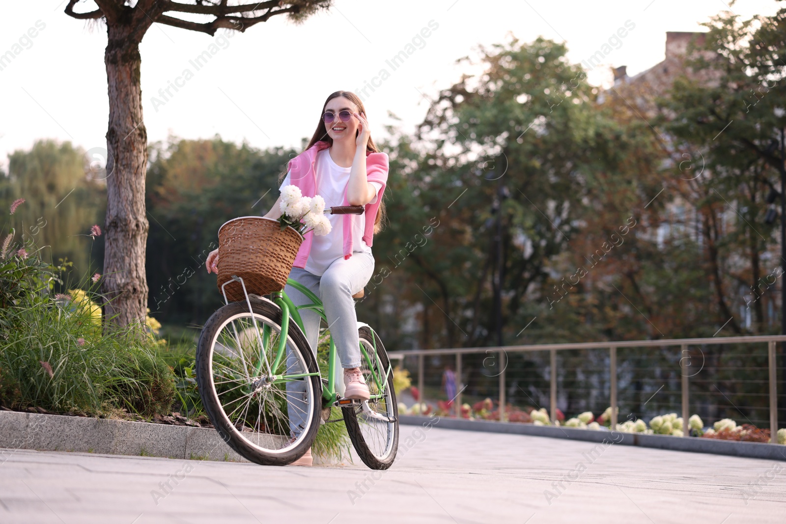 Photo of Woman riding bicycle with flowers in basket on city street. Space for text