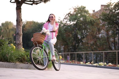 Photo of Woman riding bicycle with flowers in basket on city street. Space for text