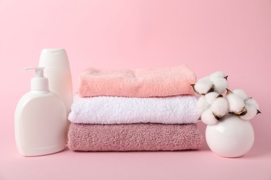 Photo of Stack of fresh terry towels, cosmetic products and cotton flowers on pink background