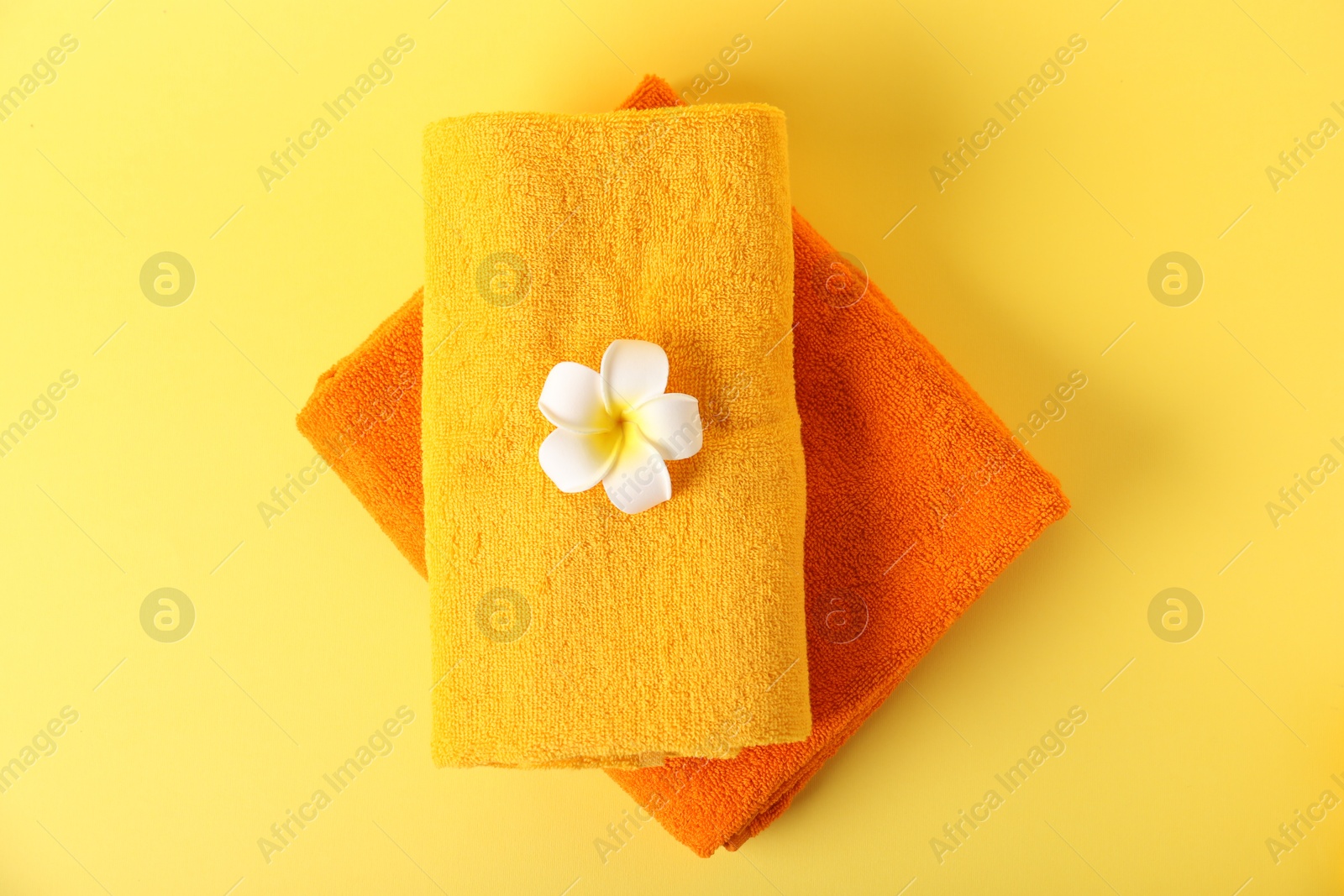 Photo of Stack of fresh terry towels and plumeria flower on yellow background, top view