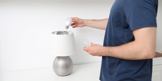 Photo of Man changing incandescent light bulb for fluorescent one in lamp indoors, closeup