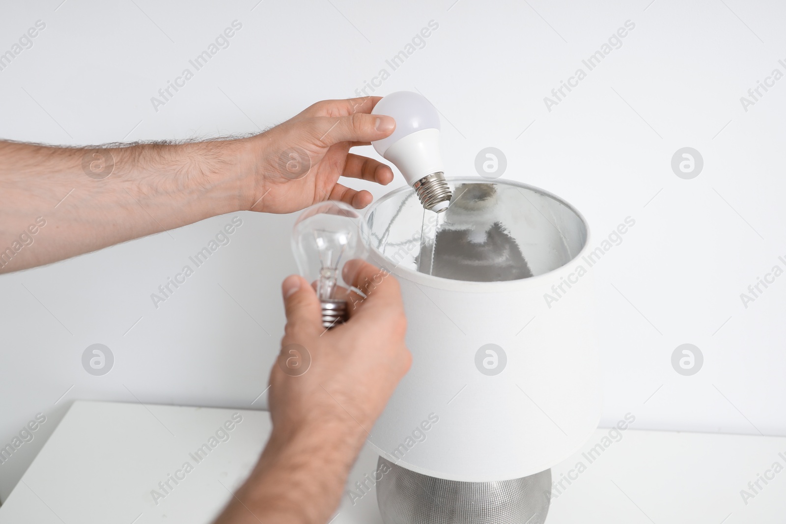 Photo of Man changing incandescent light bulb for fluorescent one in lamp on white table, closeup