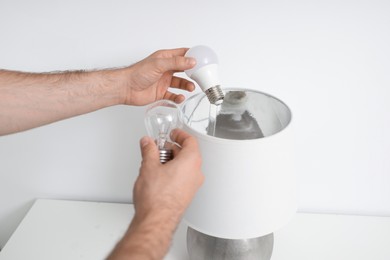 Photo of Man changing incandescent light bulb for fluorescent one in lamp on white table, closeup