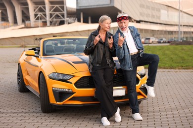 Photo of Stylish senior couple near modern cabriolet showing rock gesture outdoors