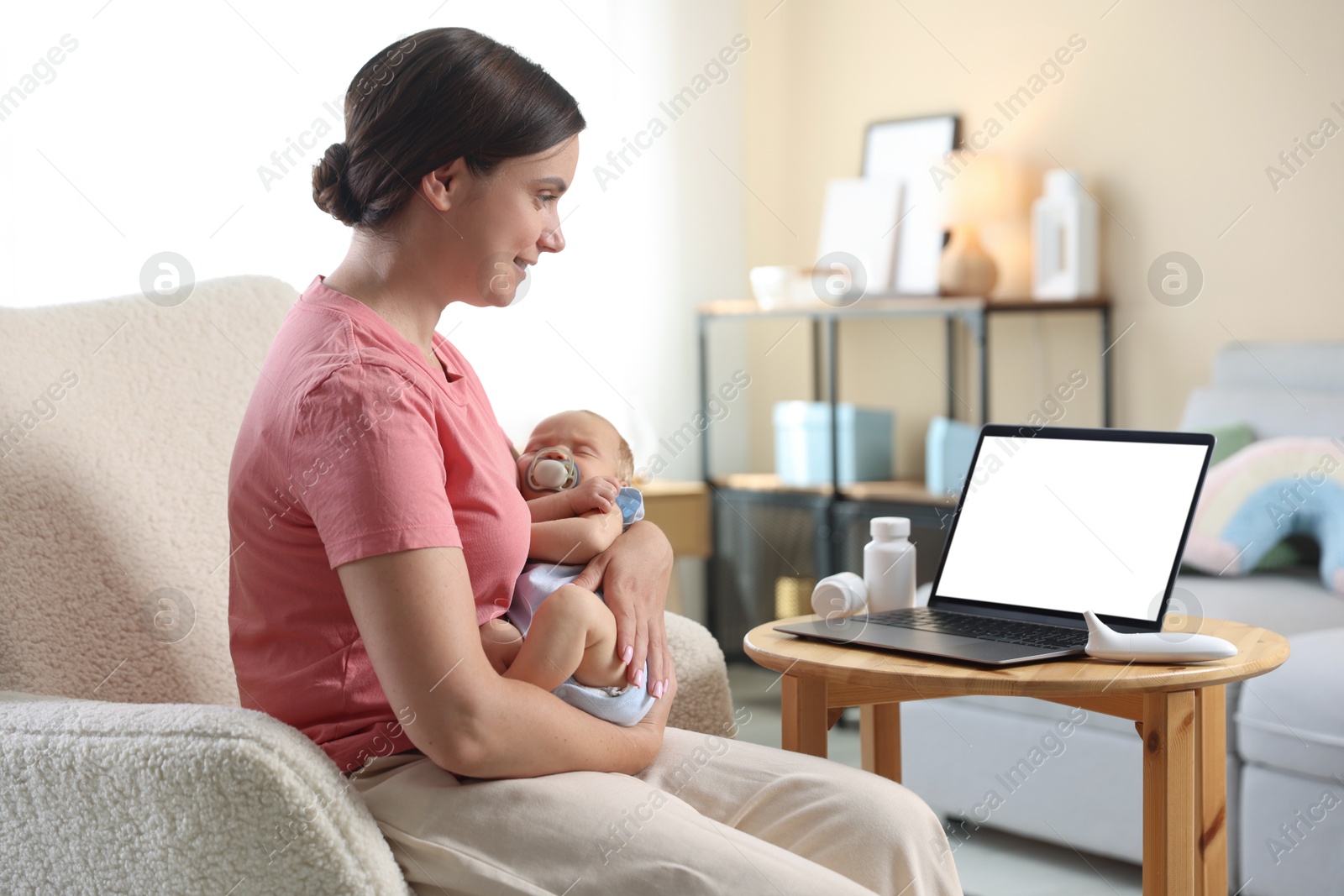 Photo of Telehealth. Smiling mother with her baby having online consultation with doctor by laptop on armchair at home