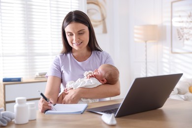 Photo of Telehealth. Smiling mother with her baby having online consultation with doctor and writing at table indoors