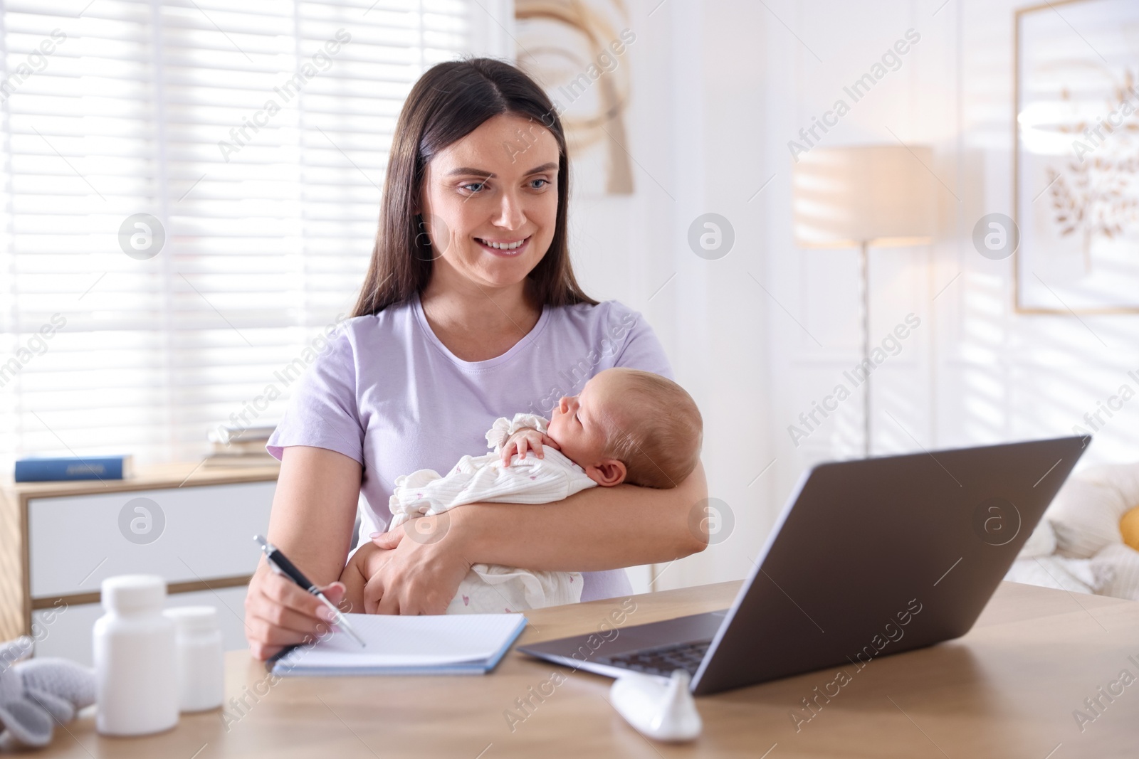 Photo of Telehealth. Smiling mother with her baby having online consultation with doctor and writing at table indoors